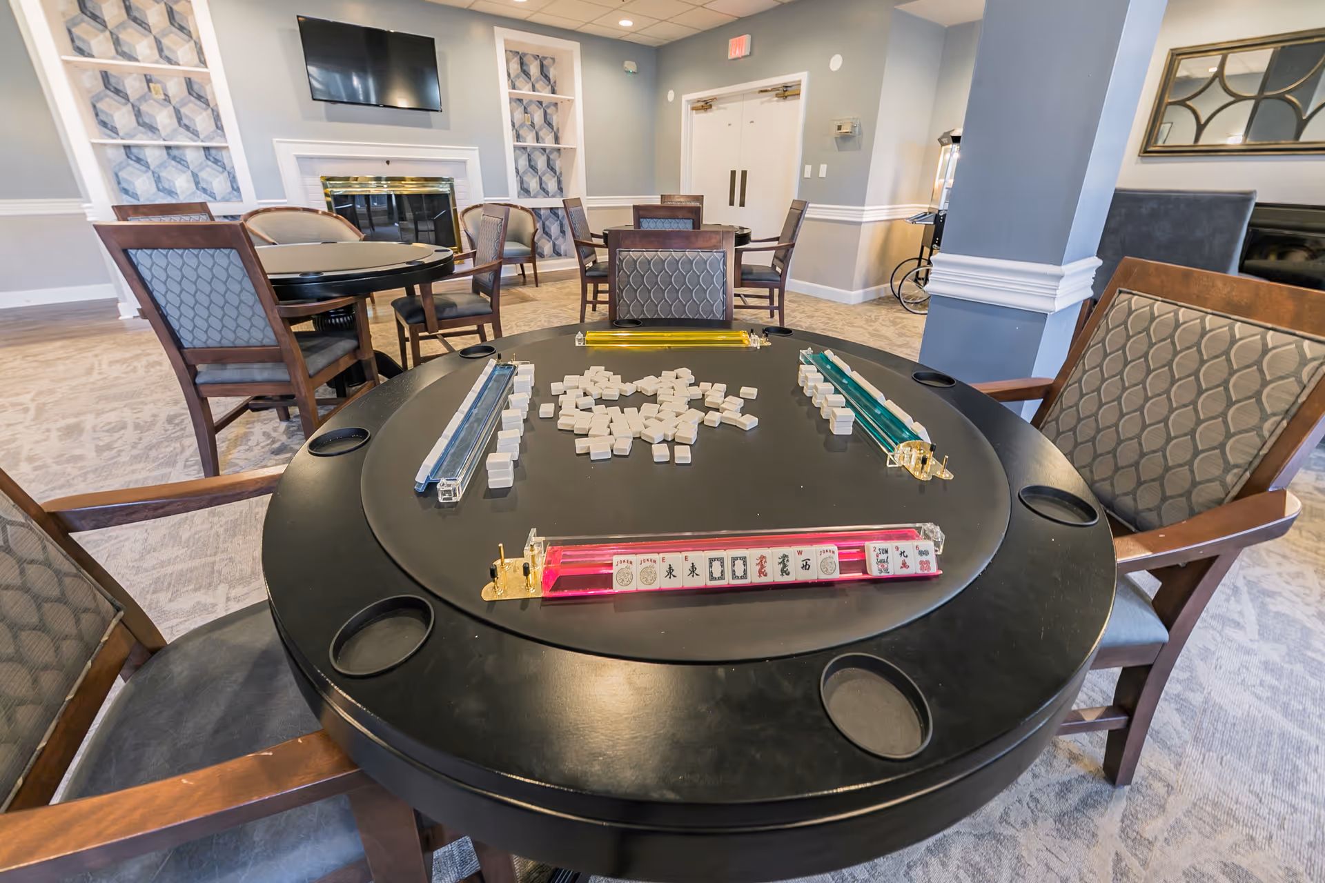 A cozy game room in a senior living facility featuring a round black table set up for a game of Mahjong with tiles and racks. The room has several wooden chairs with patterned cushions, a fireplace with a TV mounted above it, and soft blue and gray walls with decorative panels. There are additional tables and chairs in the background, and a popcorn machine is visible near a column.