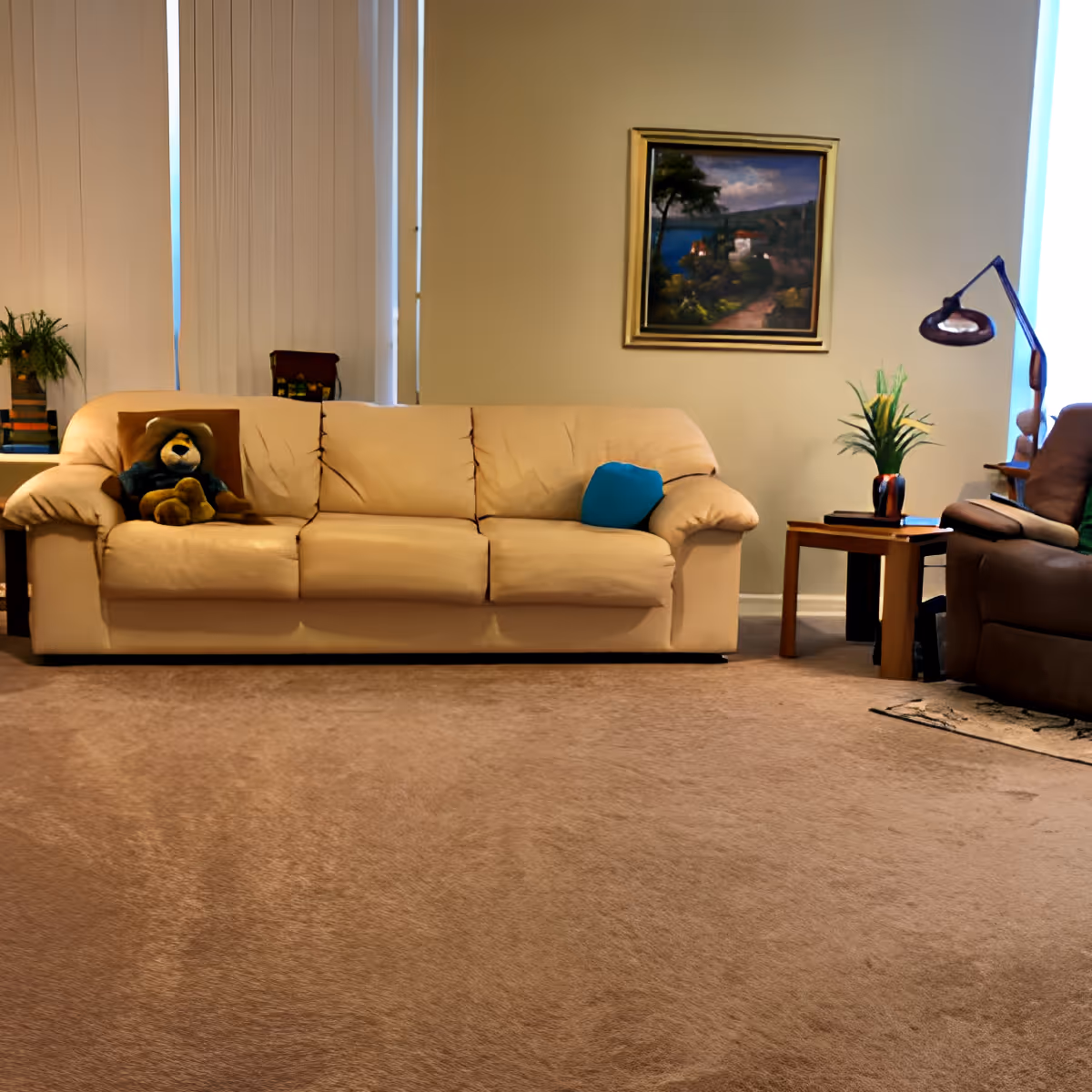 A cozy living room with a beige sofa featuring a teddy bear and a blue cushion. There is a side table with a plant and a lamp next to a brown recliner chair. A framed landscape painting hangs on the wall behind the sofa, and vertical blinds cover the windows.