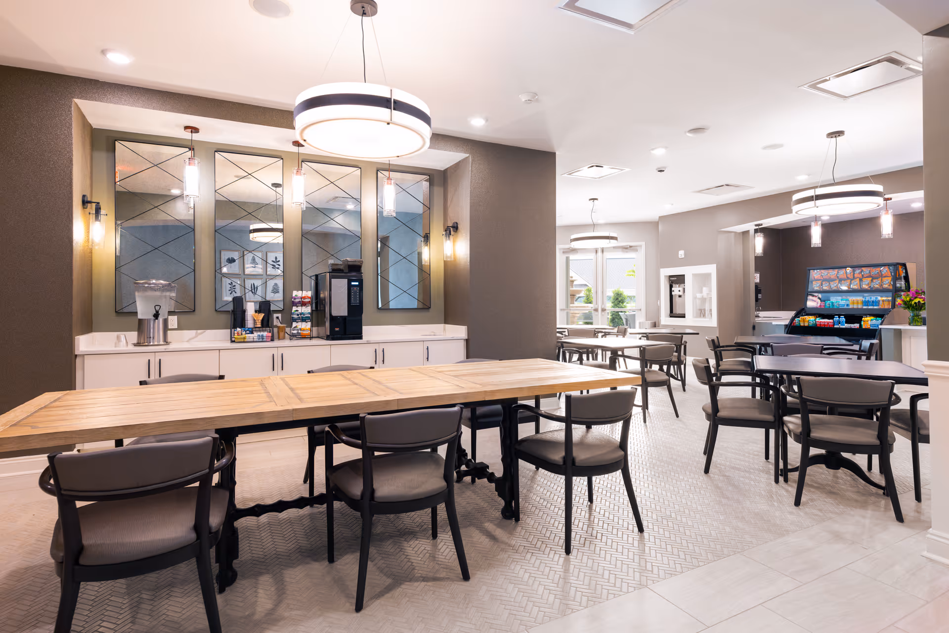A bright and modern dining area in a senior living facility featuring multiple tables and chairs arranged for communal dining. The room has contemporary light fixtures hanging from the ceiling, a beverage station with a water dispenser and coffee machine against the wall, and a snack display with various packaged items. Large mirrors and neutral-colored walls enhance the spacious and welcoming atmosphere.