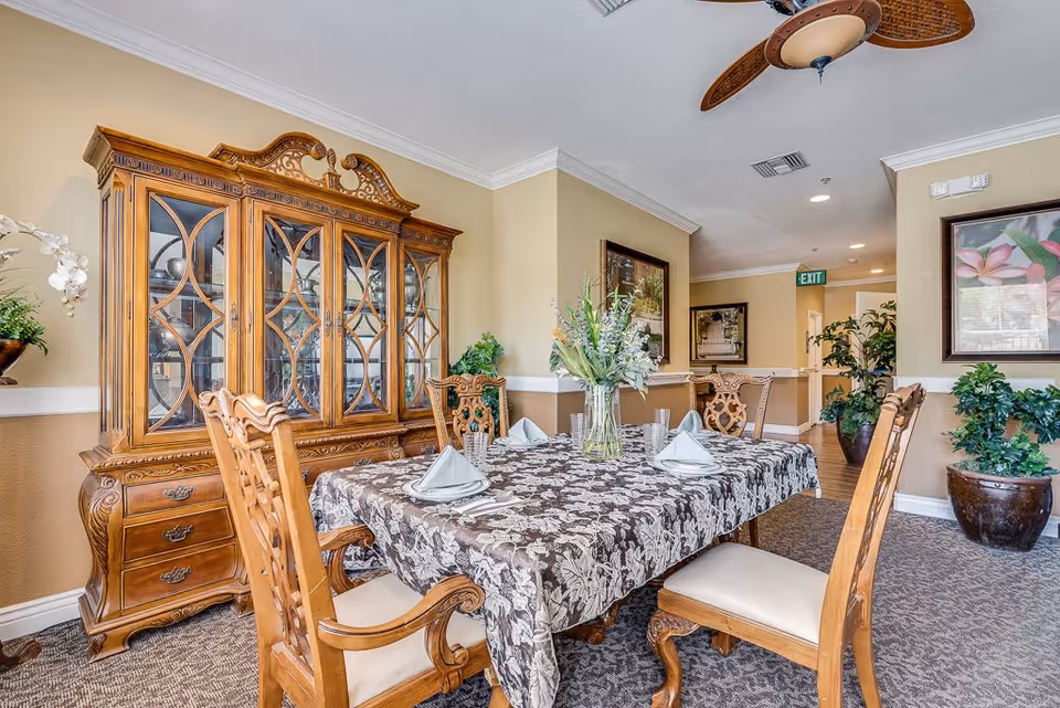A dining room with a wooden dining table covered with a floral tablecloth, set with folded napkins and glasses. Surrounding the table are ornate wooden chairs with cushioned seats. Against the wall is a large wooden china cabinet with glass doors displaying dishes. The room has beige walls, carpeted floor, ceiling fan, framed artwork, and several potted plants.