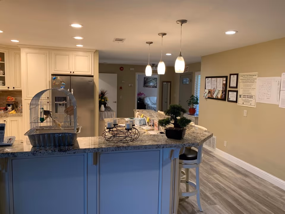 Interior view of a kitchen area in an elder care facility with a granite countertop island. On the island, there is a birdcage with a bird inside, a decorative candle holder, and a small potted plant. The kitchen has white cabinets, a stainless steel refrigerator, and three pendant lights hanging above the island. In the background, there is a hallway with a door, a bulletin board with notices on the wall, and a chair near the island.