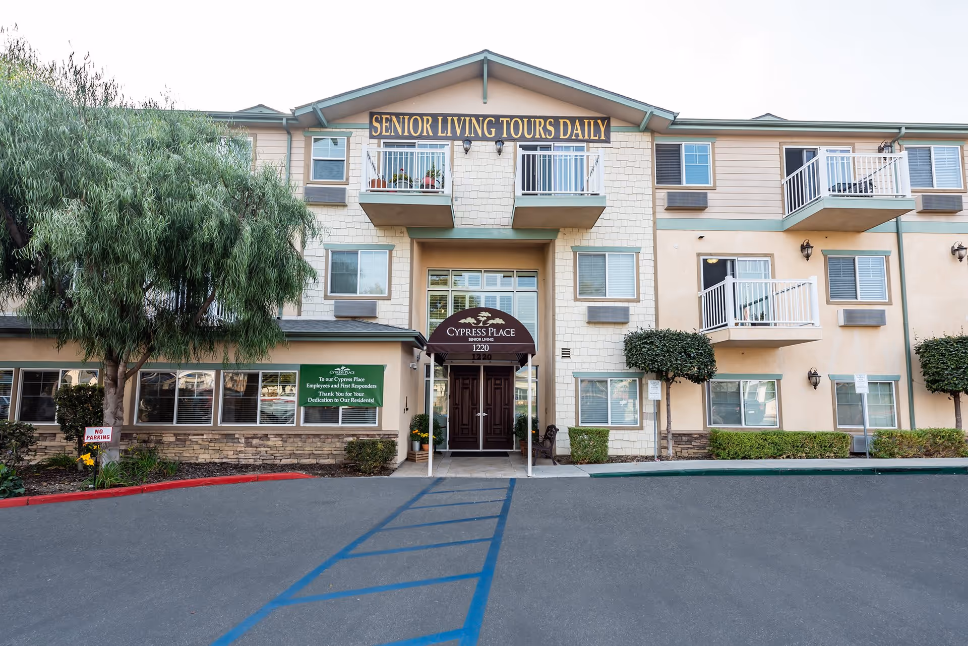 Front exterior view of Cypress Place senior living facility with a three-story building, balconies, trees, and a sign above the entrance that reads 'Senior Living Tours Daily'.