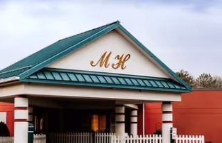 Entrance of a senior living facility with a green metal roof and white columns. The letters 'M H' are displayed prominently on the front gable above the entrance. There is a white picket fence in front of the building and some trees in the background under a cloudy sky.