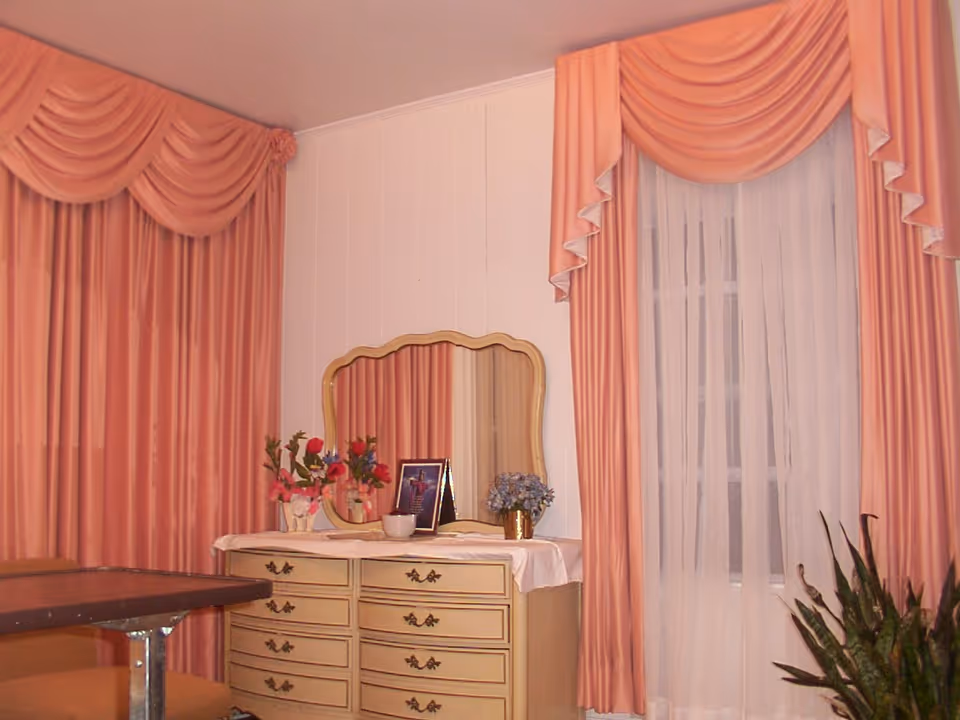 A room corner featuring a cream-colored dresser with a large ornate mirror on top. The dresser has several drawers and is decorated with flower vases and a framed picture. The walls are light-colored, and there are large windows covered with sheer white curtains and heavy peach-colored drapes with valances. A plant is visible in the bottom right corner.