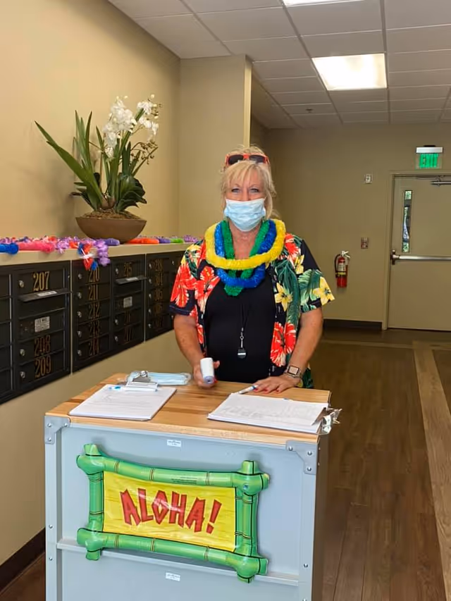 A woman wearing a face mask and colorful Hawaiian-themed clothing and leis stands behind a small desk with clipboards and papers. The desk has a sign that says 'ALOHA!' and is located in a hallway with mailboxes on the wall and a potted plant on top of the mailboxes.
