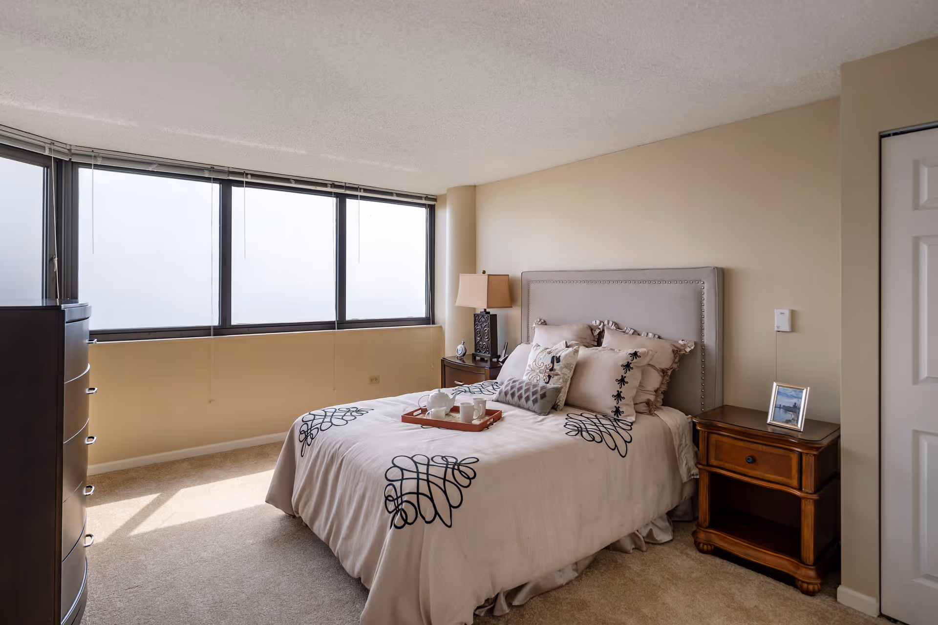 A bedroom with a large window letting in natural light. The room features a bed with a light-colored headboard and bedding decorated with black patterns. There is a wooden nightstand with a lamp and a small clock on one side of the bed, and another wooden nightstand with a framed photo on the other side. A dark wooden dresser is positioned near the window.