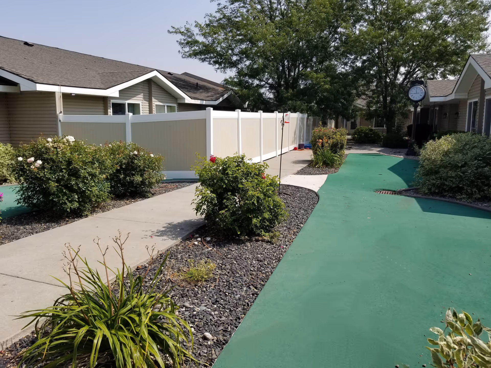 Outdoor pathway at Gables of Ammon Assisted Living with a green walking path and concrete sidewalk bordered by bushes and plants. Single-story buildings with beige siding and white fences are visible on both sides. A black street clock is seen in the distance under a large tree.