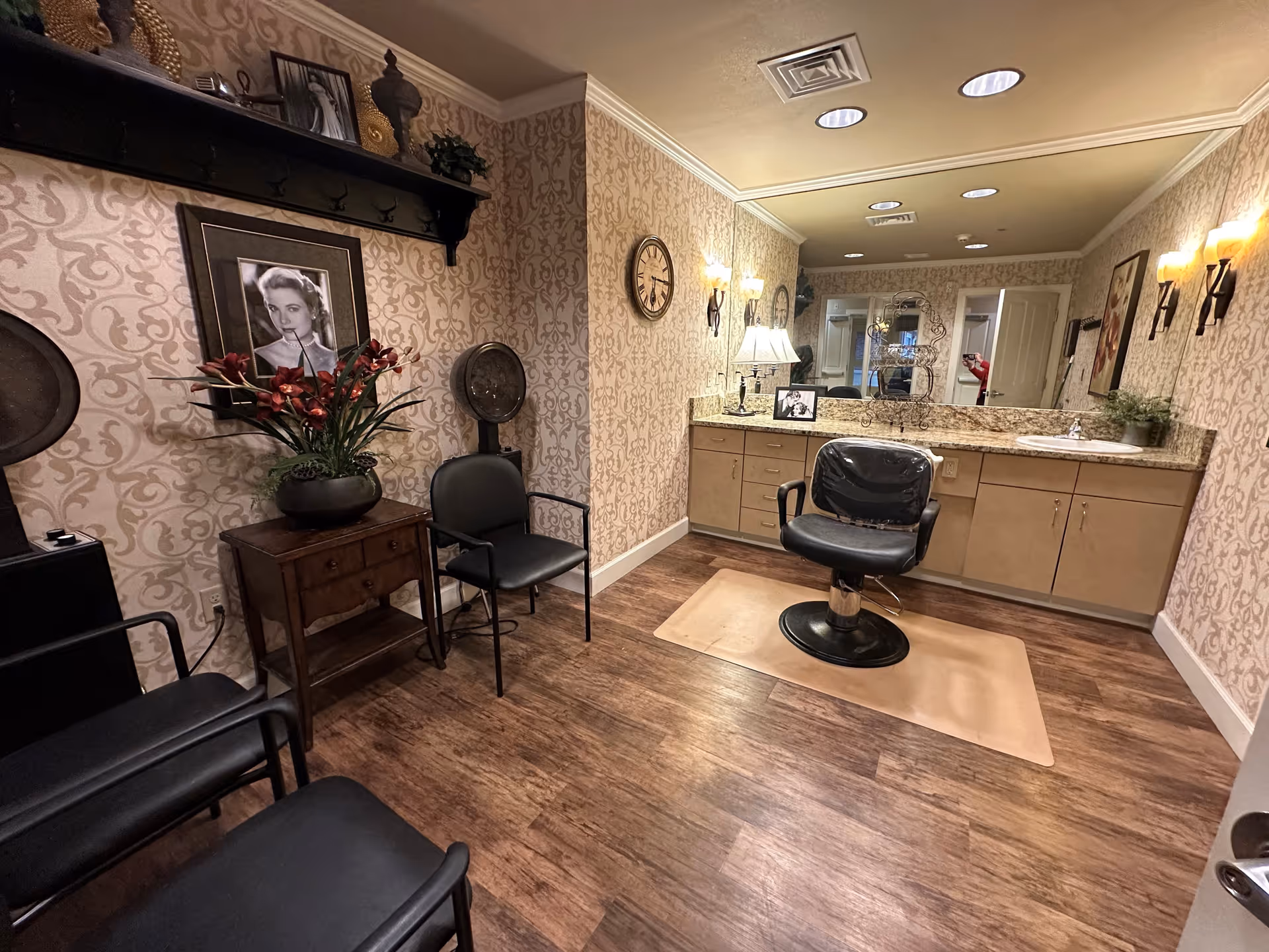 Interior view of a hair salon area in a senior living facility with a styling chair in front of a large mirror and countertop with cabinets. The room has patterned wallpaper, wooden flooring, wall-mounted lights, a clock, framed black and white photos, and several black chairs along the wall. A small table with a plant and decorative items is also visible.