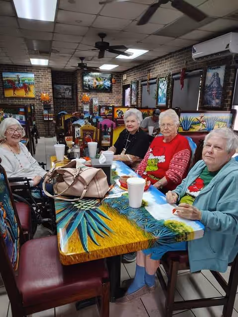 Four elderly women sitting around a colorful painted table in a dining area with brick walls and vibrant artwork. They have drinks in white cups and appear to be enjoying each other's company.