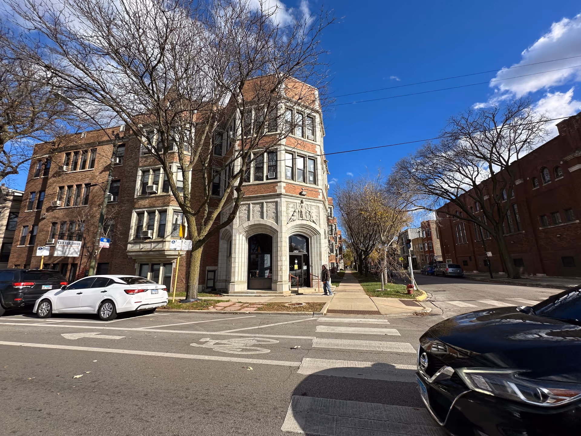 Street view of a multi-story brick building with ornate stone detailing at the corner entrance, labeled Center Home for Hispanic Elderly. Several cars are parked along the street, leafless trees line the sidewalk, and the sky is clear with some clouds.