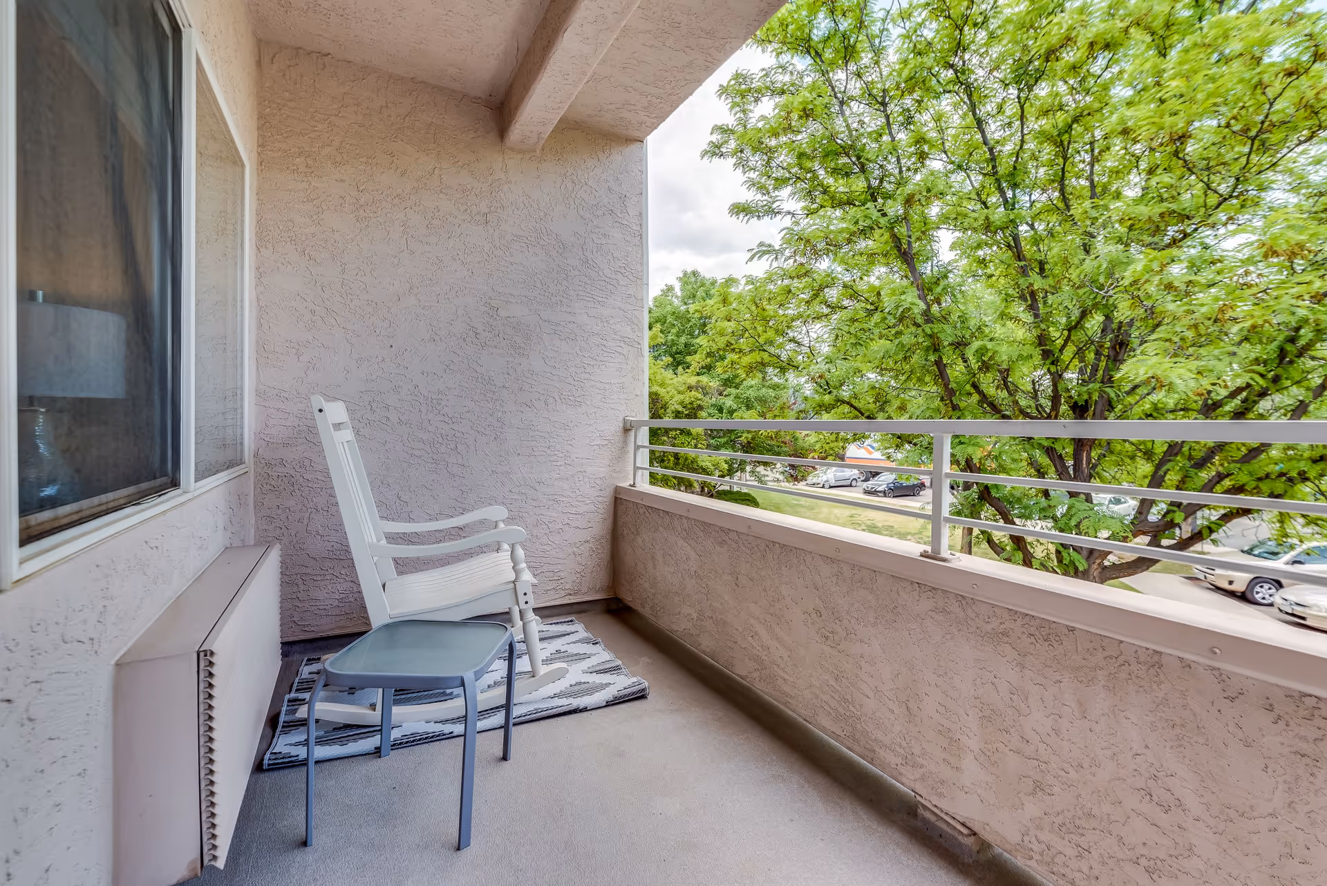 A small outdoor balcony with a white rocking chair and a small gray side table on a patterned rug. The balcony has textured beige walls and a metal railing overlooking green trees and parked cars.