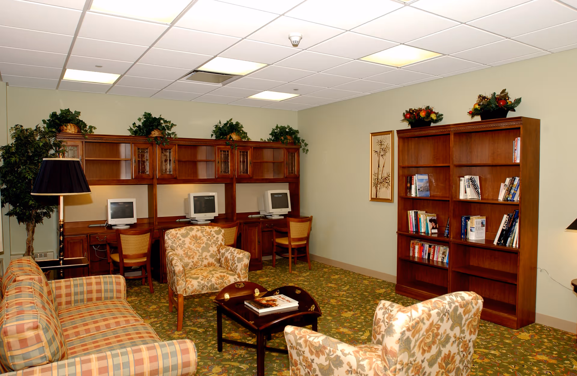 A cozy common area with a plaid sofa, two floral armchairs, a small wooden coffee table with books, a wooden bookshelf filled with books, and a row of desks with vintage computer monitors. The room has a green patterned carpet, light green walls, and ceiling lights. There are plants and decorative items on top of the furniture.