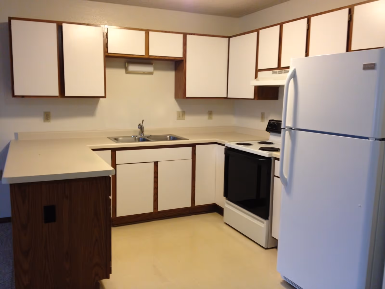 A kitchen with white countertops and cabinets with wood trim, a double stainless steel sink, a white electric stove with four burners, a white refrigerator, and beige flooring.