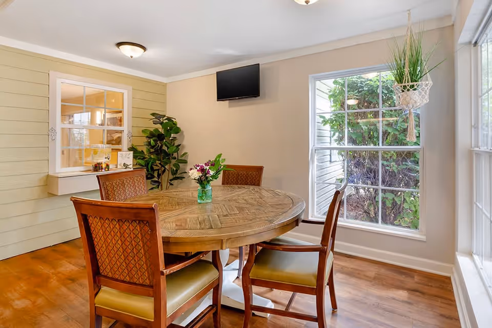 A cozy dining area with a round wooden table surrounded by four wooden chairs with cushioned seats. A small vase with flowers is placed in the center of the table. The room has large windows allowing natural light to enter, a potted plant in the corner, and a wall-mounted flat-screen TV. The walls are painted in light colors, and the floor is wooden.