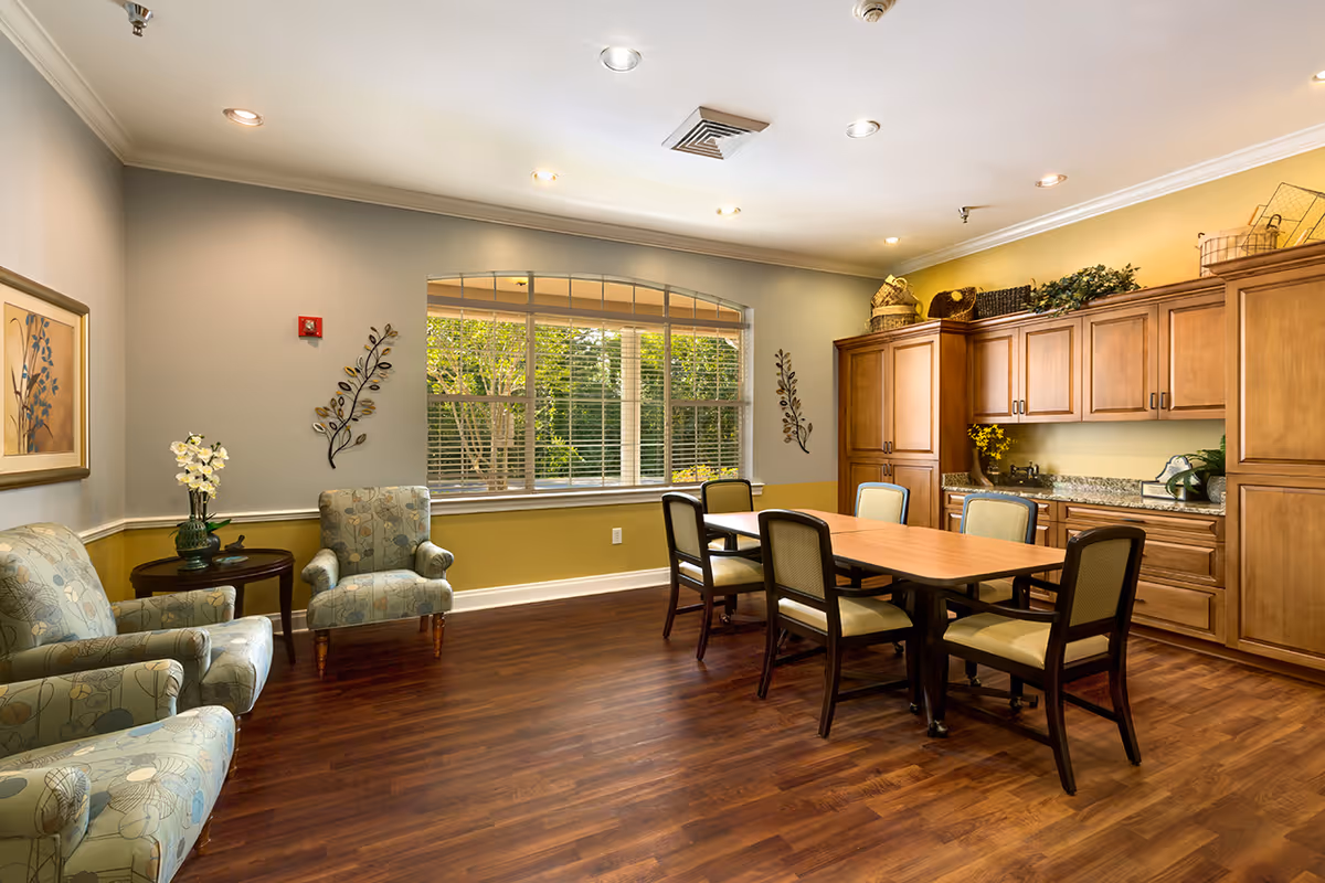 A well-lit room with wooden flooring featuring a rectangular wooden table surrounded by six cushioned chairs. To the right, there are wooden cabinets with decorative baskets and plants on top. On the left side, there are two patterned armchairs with a small round table between them holding a vase with white flowers. A large window with blinds lets in natural light, and the walls are painted in soft gray and yellow tones with decorative wall art.