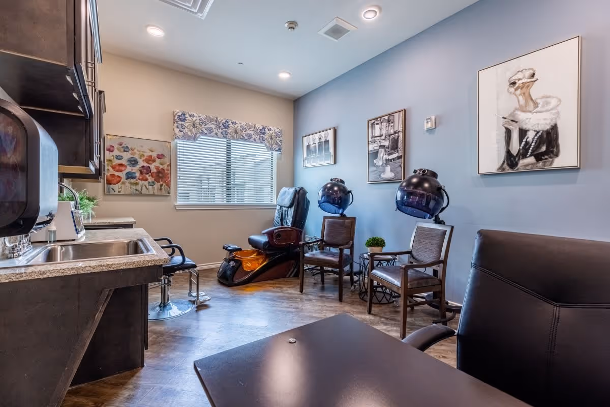 Interior view of a salon room with two black hair drying chairs, two wooden chairs, a pedicure chair with a foot bath, a countertop with a sink, and wall art including a painting of a woman and floral artwork. The room has a window with blinds and a floral valance.