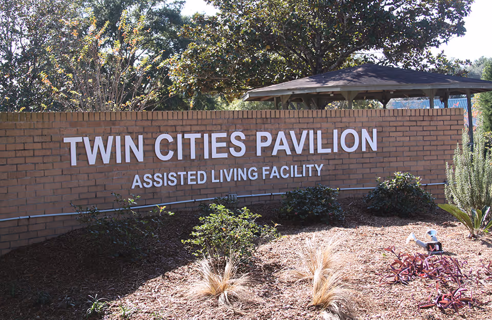 Brick entrance sign reading 'Twin Cities Pavilion Assisted Living Facility' set amid landscaping with a pavilion and trees behind.