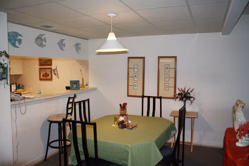 Small dining area with a green-tablecloth covered table, black chairs, pendant light, and a kitchen pass-through with decorative items.