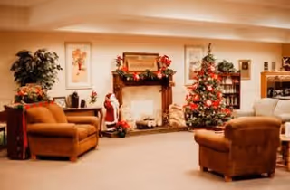 A cozy living room decorated for Christmas with a Christmas tree, garlands on the fireplace mantel, and festive ornaments. The room has comfortable brown armchairs, framed artwork on the walls, and bookshelves filled with books.