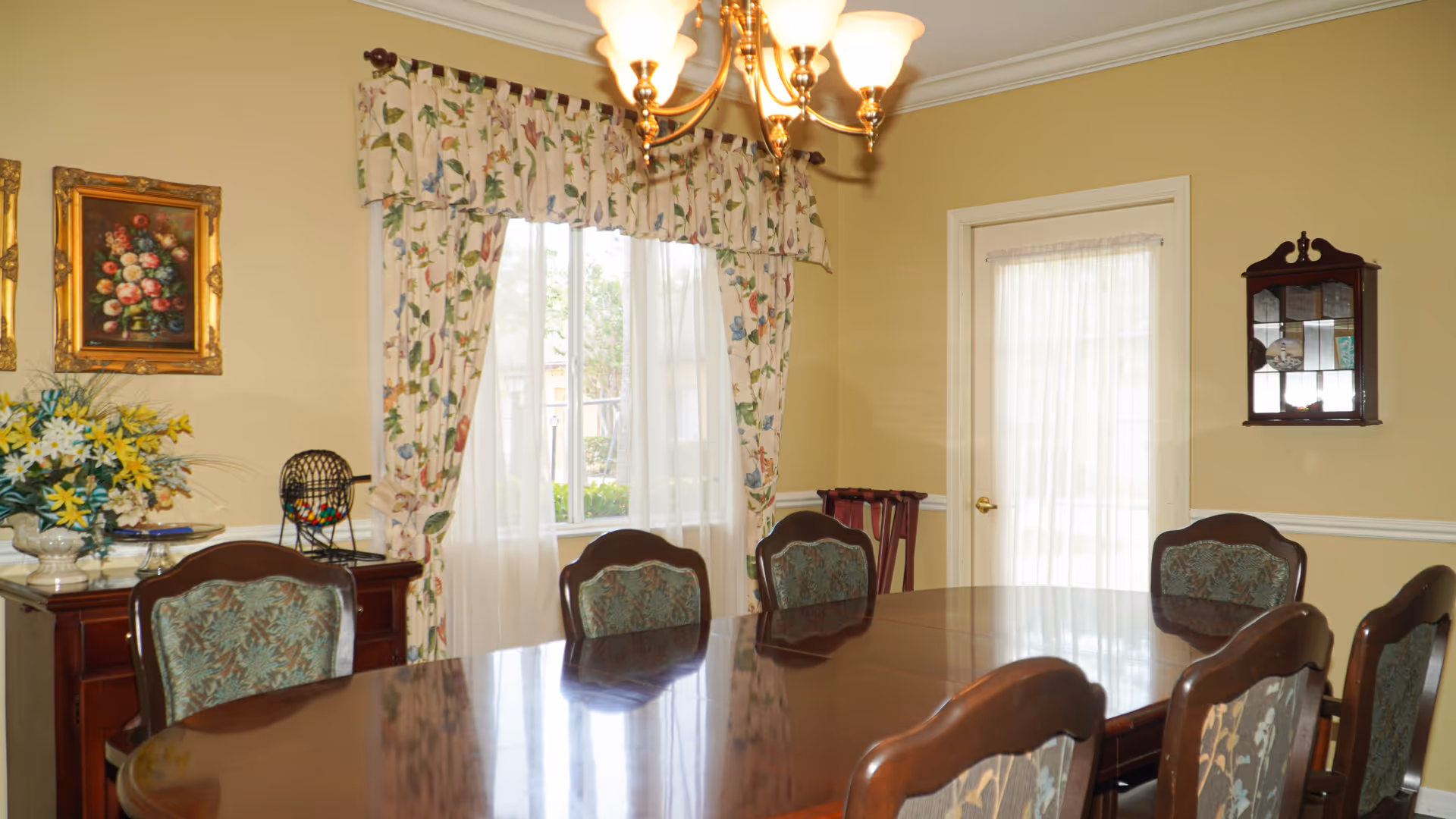 Formal dining room with a long polished wooden table surrounded by upholstered chairs, a chandelier, floral curtains, and a sideboard with flowers.