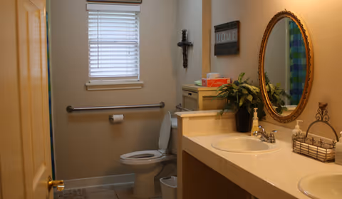 A small assisted-living bathroom with a toilet and grab bar, double sink vanity, oval mirror, and decorative wall accents.
