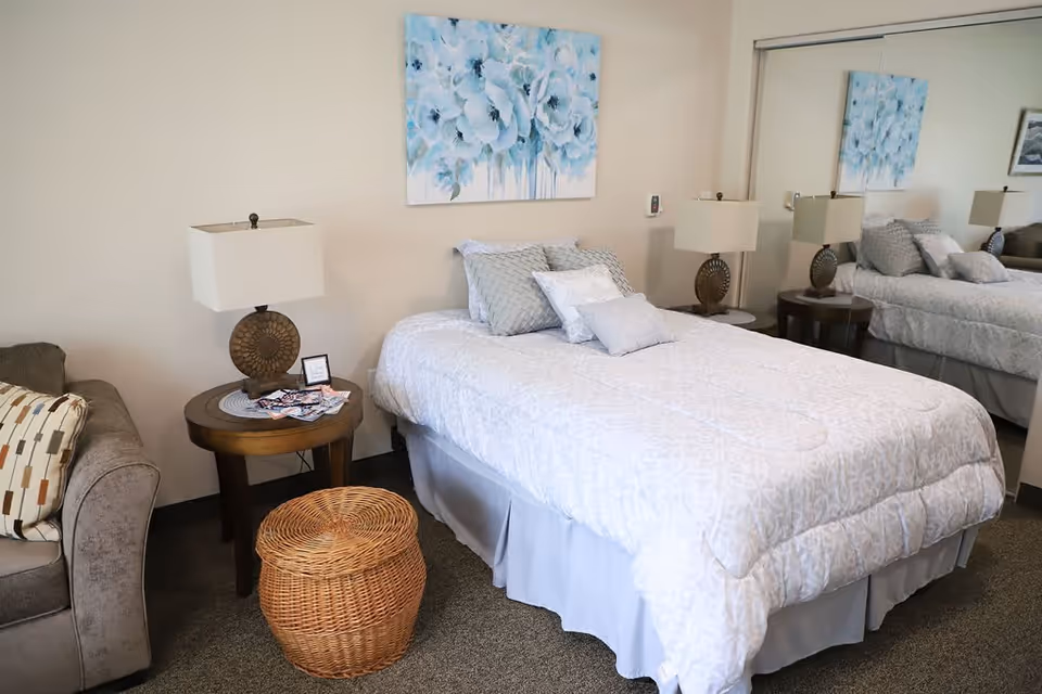 A neatly made bed with white and light gray patterned bedding and multiple pillows in a bedroom. On either side of the bed are round wooden nightstands with matching lamps. A wicker basket is placed at the foot of the bed. A large mirror covers the wall behind one side of the bed, reflecting the room. A couch with a patterned pillow is partially visible on the left side. Above the bed hangs a painting of blue flowers.