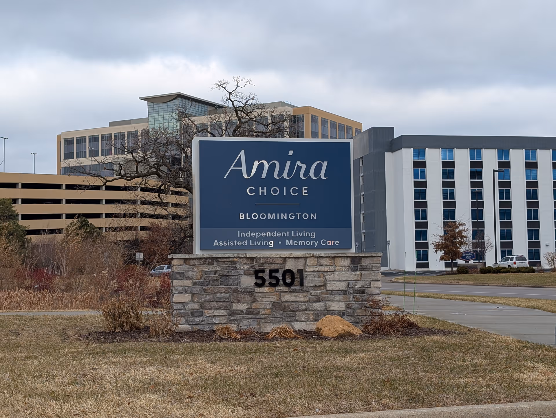 Outdoor view of a stone and blue sign for Amira Choice Bloomington, an independent living, assisted living, and memory care facility, with buildings and a parking structure in the background under a cloudy sky.