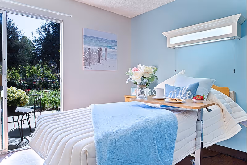 A bright and cozy bedroom with a hospital-style adjustable bed covered in white and light blue bedding. A wooden overbed table holds a plate with food, a cup, and a bowl of strawberries. There is a decorative pillow with the word 'smile' on the bed. A vase with flowers is on a bedside table next to the bed. The room has light blue walls and a large open door leading to an outdoor patio with a small table and chairs surrounded by greenery.