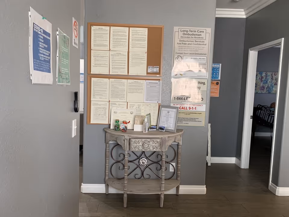 Interior view of a hallway corner in an elderly care facility with gray walls and a small decorative wooden table holding hand sanitizer and framed certificates. Several informational posters and notices, including COVID-19 guidelines and Long-Term Care Ombudsman information, are pinned on the walls. Doorways lead to other rooms, one of which shows a bed with floral bedding.