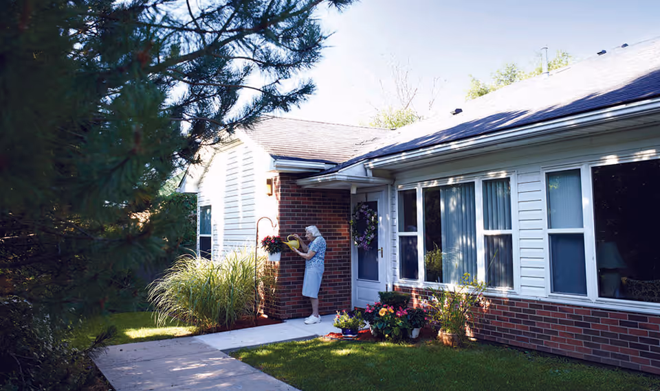 An elderly woman watering hanging plants outside a single-story brick and siding building with large windows, surrounded by green grass and trees on a sunny day.