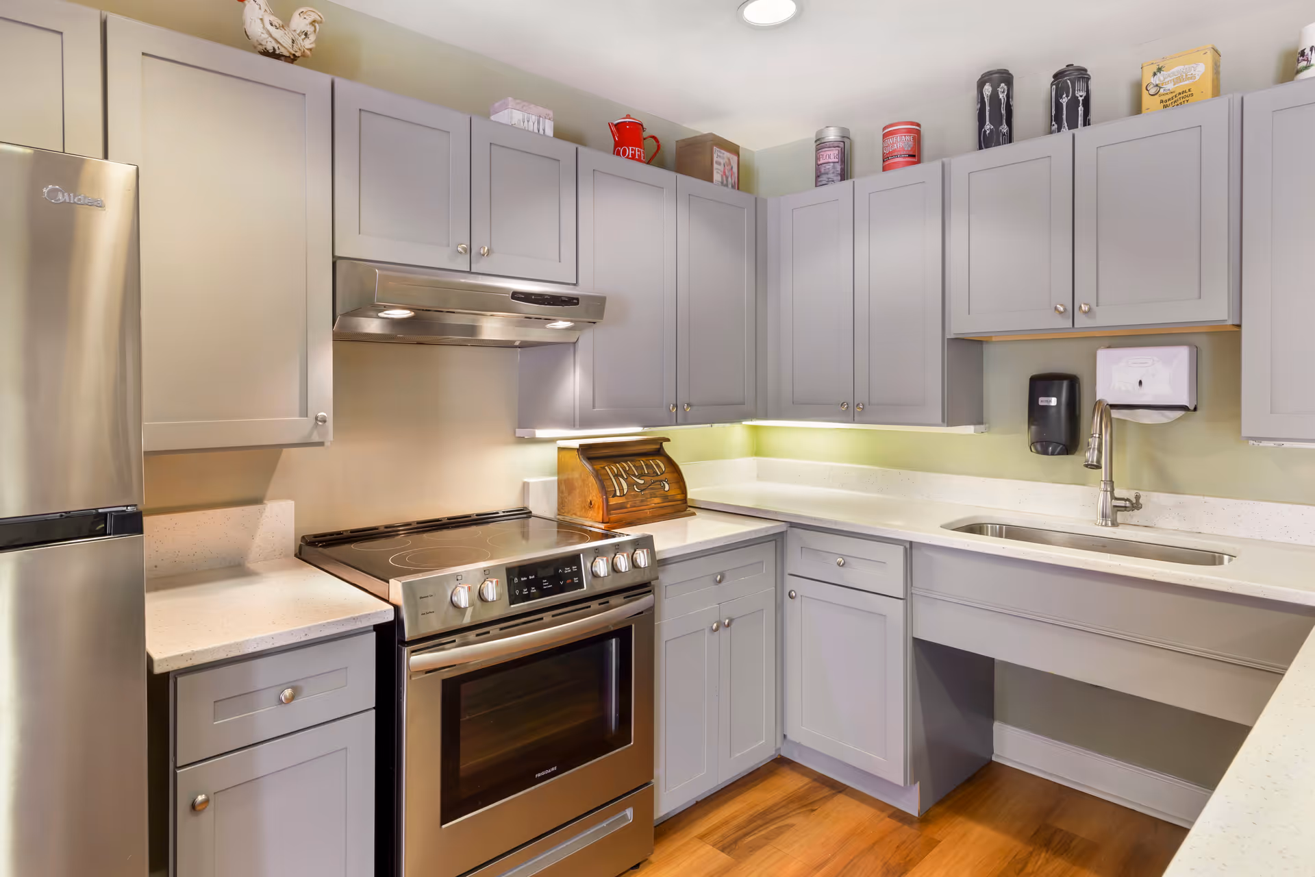 A modern kitchen with light gray cabinets, a stainless steel refrigerator, an electric stove with an oven, a stainless steel range hood, and a sink with a faucet. The countertops are white, and there are various decorative items on top of the cabinets. The floor is wooden.