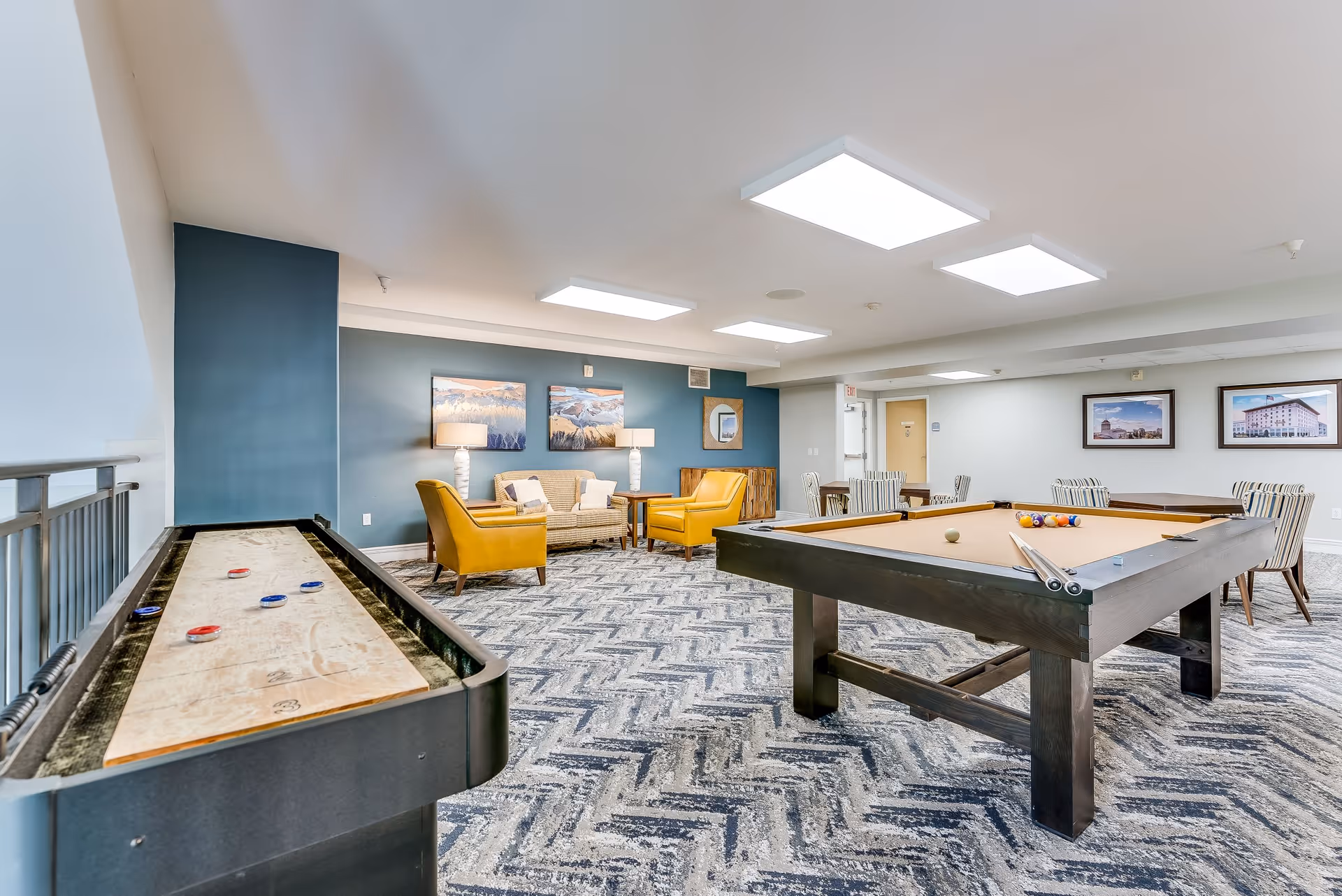 A recreational room in a senior living facility featuring a shuffleboard table on the left and a pool table on the right. The room has patterned carpet flooring, a seating area with two yellow armchairs and a beige sofa against a blue accent wall with two landscape paintings and two table lamps. There are also several tables with striped chairs along the back wall, and bright ceiling lights illuminate the space.
