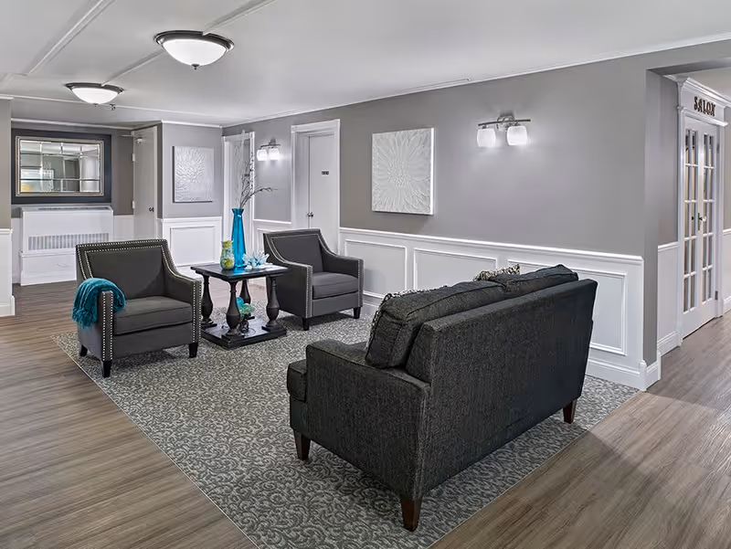 A cozy senior living facility common area with two dark gray armchairs and a matching sofa arranged around a small wooden table with decorative vases. The room features gray walls with white wainscoting, a patterned area rug, and soft lighting from ceiling fixtures and wall sconces. A door labeled 'Salon' is visible on the right side.