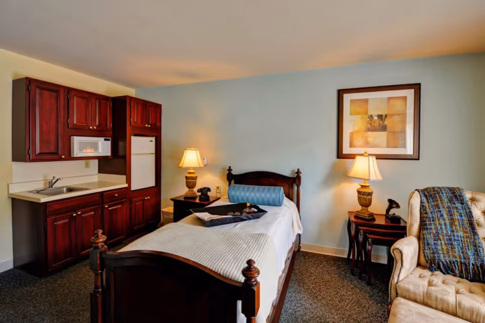 Furnished senior living bedroom with a single bed, bedside tables and lamps, a small kitchenette, and an armchair.