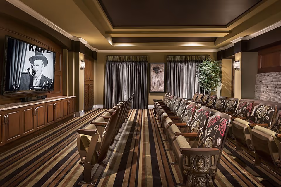 A cozy theater room with two rows of patterned upholstered chairs facing a large screen displaying a black and white movie scene. The room features striped carpet, warm lighting, wood paneling, and curtains covering the windows at the back.