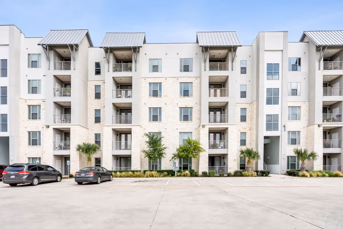 Exterior view of a modern multi-story senior living facility building with balconies, windows, and a parking lot in front with a few parked cars. The building has light-colored stone and stucco facade with metal awnings above some balconies.