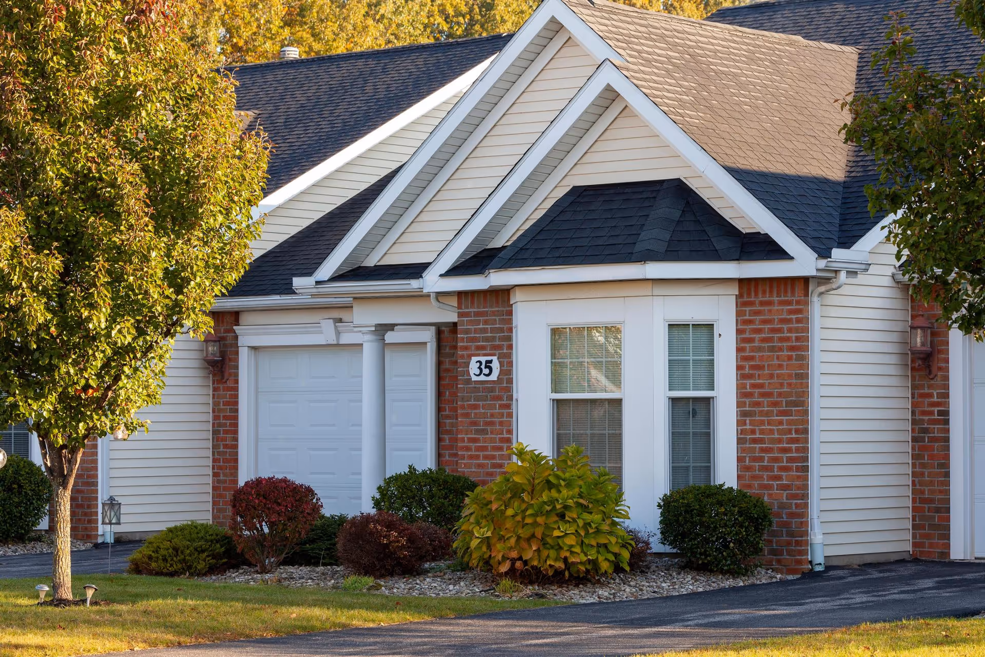 Exterior view of a residential building with a white garage door, brick and beige siding, a bay window, and a driveway. There are small bushes and a tree with green and yellow leaves in front of the building.