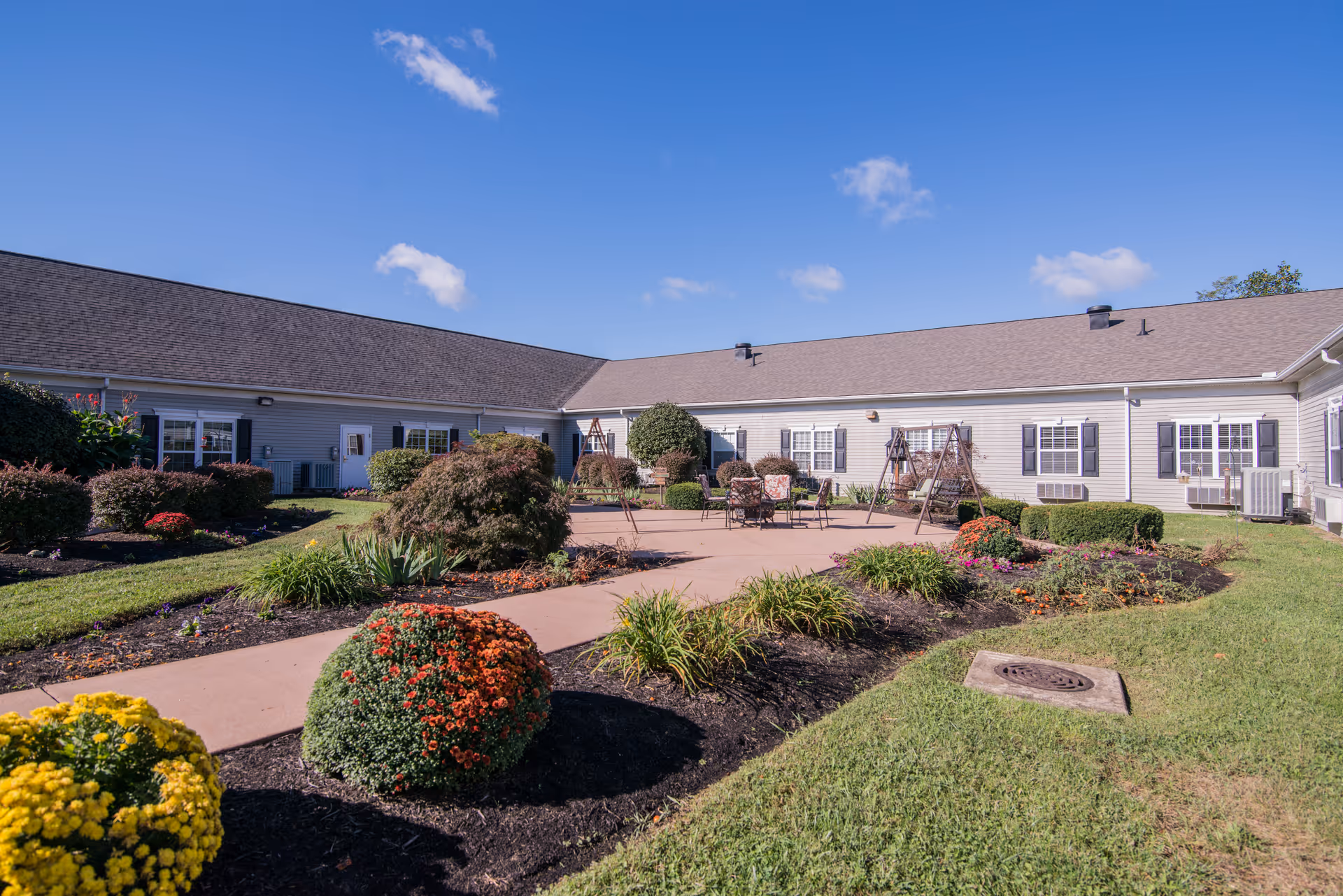 Outdoor courtyard area of a senior living facility with a paved walkway, landscaped flower beds with colorful flowers and shrubs, patio furniture, and swing seats. The building surrounds the courtyard with multiple windows and a clear blue sky overhead.