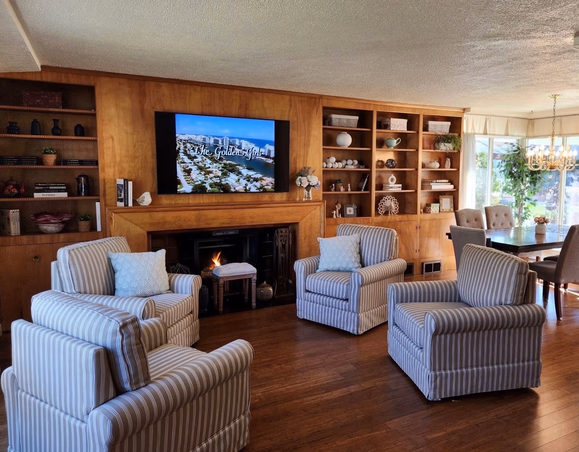 A cozy living room with four striped armchairs arranged around a fireplace. Above the fireplace is a mounted flat-screen TV displaying the title 'The Golden Girls.' The room features wooden built-in shelves filled with decorative items and books. To the right, there is a dining area with a table, chairs, a chandelier, and large windows letting in natural light.