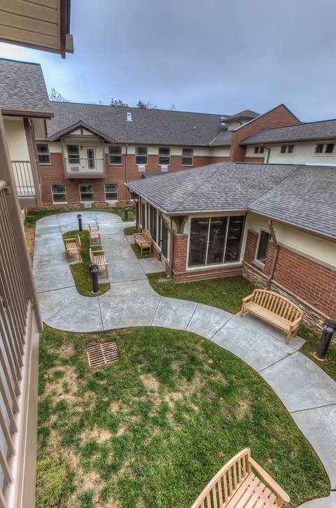 View of an outdoor courtyard area at a senior living facility with concrete walkways, several wooden benches, green grass patches, and a two-story brick building surrounding the courtyard under a cloudy sky.