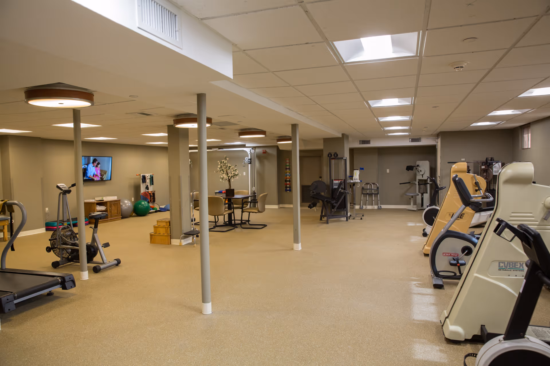 A spacious fitness and rehabilitation room with various exercise equipment including stationary bikes, a treadmill, and weight machines. There is a small table with chairs in the center, a TV mounted on the wall, and exercise balls and other therapy tools in the background. The room has beige walls and ceiling tiles with bright lighting.