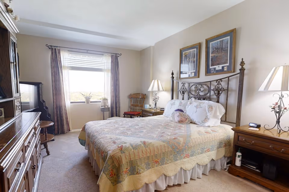 Sunlit bedroom with a decorative metal bed topped with a quilt, wooden nightstands and dresser, and a window with curtains.