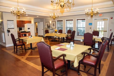 A dining room with several tables covered in yellow tablecloths and set with placemats, napkins, and condiments. The room has wooden floors, large windows with blinds, and multiple brass chandeliers hanging from the ceiling. There is a small kitchen area with a refrigerator and microwave in the background.