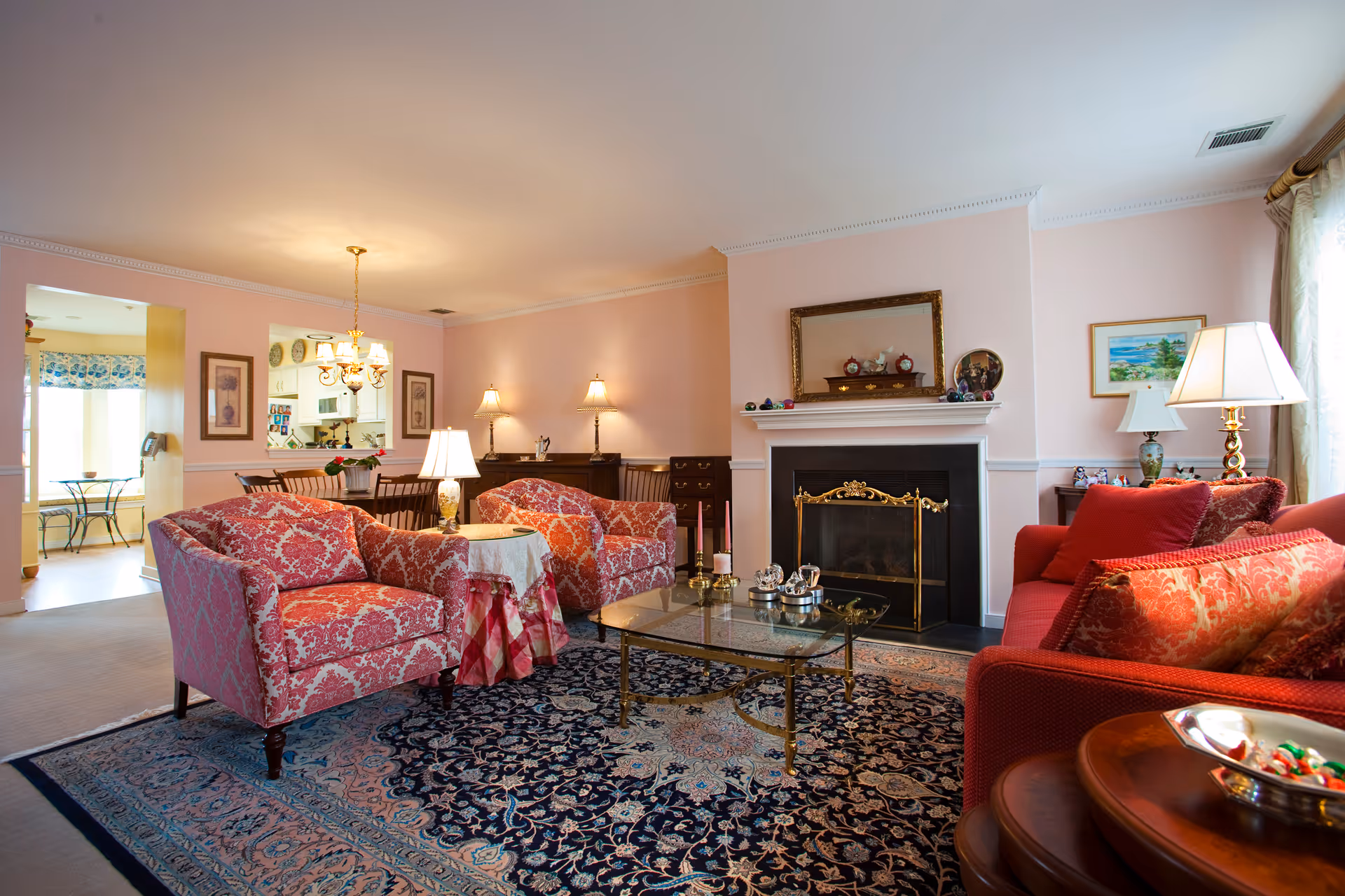 A cozy living room with a red patterned sofa and two matching armchairs arranged around a glass coffee table on a large ornate rug. The room features a fireplace with a gold screen and a mirror above it, two table lamps on side tables, and a dining area with a chandelier and wooden chairs in the background. The walls are painted light pink and decorated with framed artwork.
