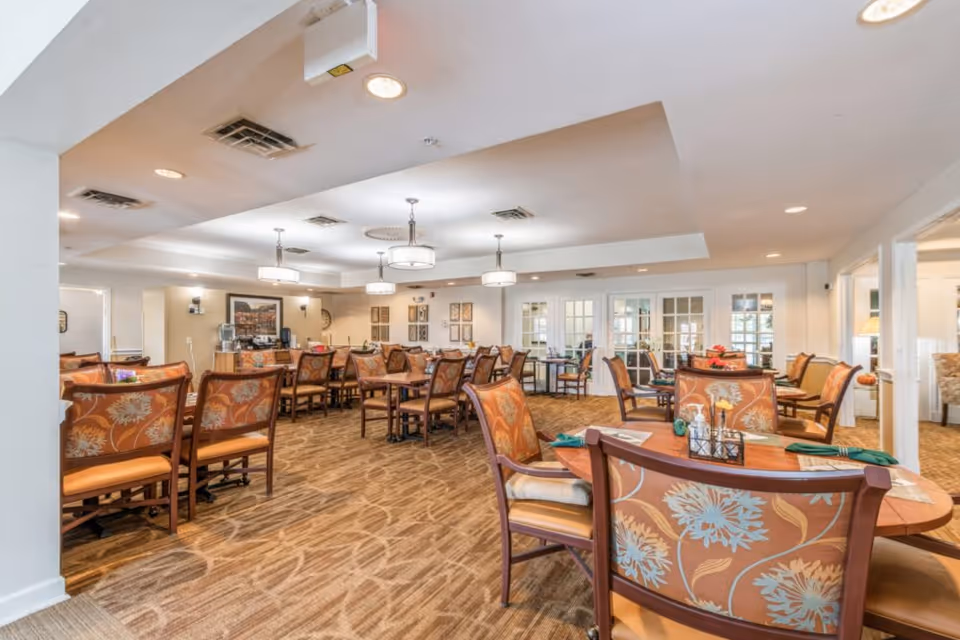 A spacious dining room in Jefferson Manor with multiple wooden tables and chairs featuring floral upholstery. The room is well-lit with ceiling lights and has a carpeted floor. There are decorative wall hangings and a water dispenser visible in the background.