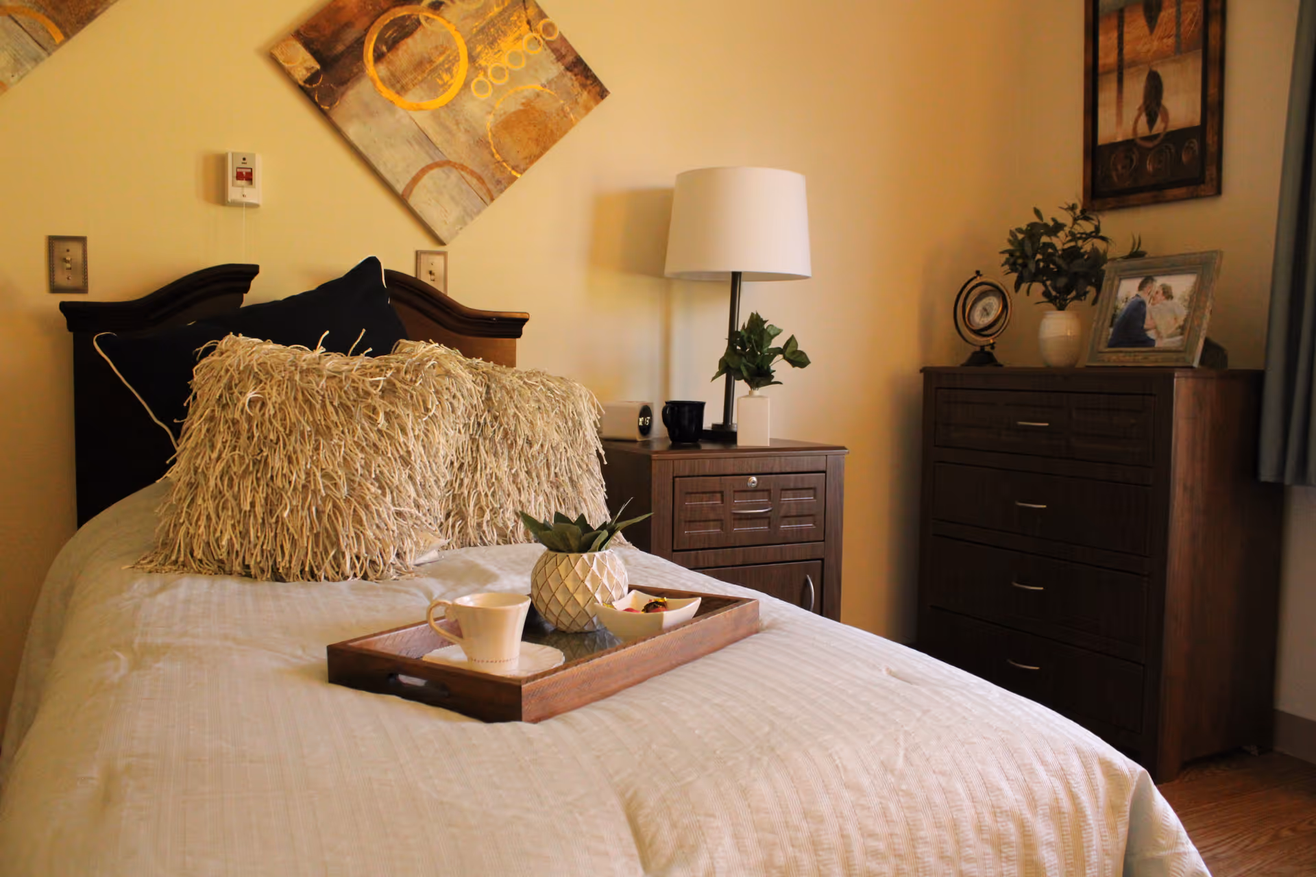 A cozy bedroom with a neatly made bed featuring textured pillows and a wooden tray holding a cup, a small plant, and a bowl. Next to the bed is a wooden nightstand with a lamp and a small plant. In the background, there is a wooden dresser with decorative items including a framed photo, a small globe, and a plant. The walls are decorated with abstract artwork.