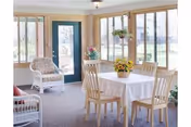 A bright and cozy room with large windows letting in natural light. The room features a round table covered with a white tablecloth and surrounded by four wooden chairs. A vase with yellow flowers is placed on the table. There are two white wicker chairs with cushions near the windows and a blue door leading outside. The room has a carpeted floor and light-colored walls.