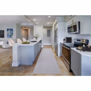 Modern kitchen with a long island countertop featuring a sink and decorative items. Stainless steel appliances including a refrigerator, microwave, and stove are visible. The kitchen opens into a living area with chairs and a painting on the wall.