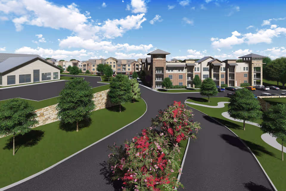 Exterior view of a senior living facility named Watermere at the Preserve, showing multiple three-story buildings with brick and light-colored siding, surrounded by landscaped greenery, trees, and a curved driveway with flowering bushes in the median under a partly cloudy blue sky.