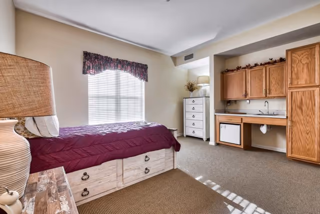 A senior living facility bedroom with a single bed covered in a purple quilt, a wooden nightstand with a lamp, a window with floral valance, a white chest of drawers with a lamp and plant on top, and a kitchenette area with wooden cabinets, a sink, and a mini refrigerator.